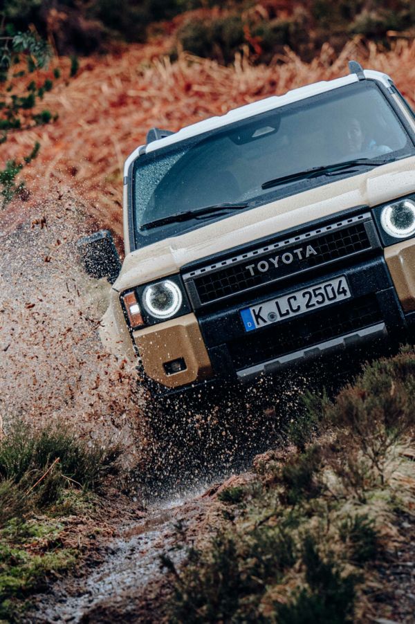 Ein beiger Toyota Landcruiser, der durch schlammiges Gelände fährt mit grünem Laub im Hintergrund.