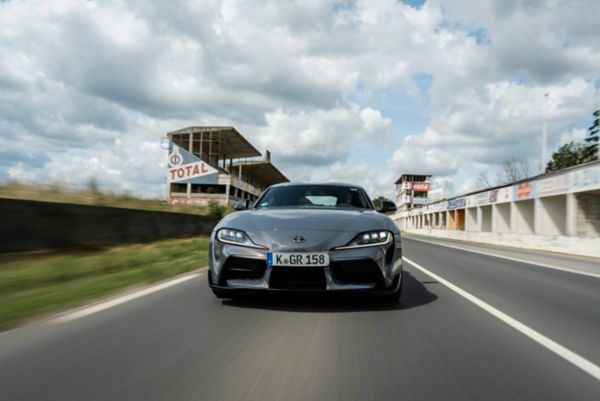 Ein grauer GR Supra von vorn fotografiert auf einer leeren Rennstrecke fahrend, im Hintergrund eine alte Tribüne und die Boxengasse mit Werbetafeln