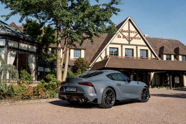 Ein grauer GR Supra von schräg hinten fotografiert vor einem alten französischen Landgasthaus im Sonnenschein