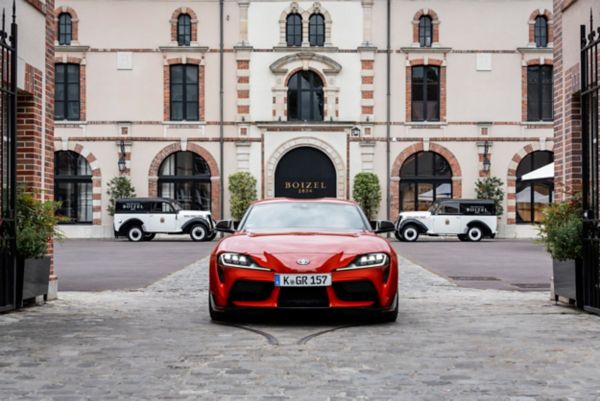 Ein roter GR Supra von vorn fotografiert vor einem Gebäude von Boizel, symmetrisch geparkt im Hintergrund zwei schwarz-weiße Oldtimer von Boizel