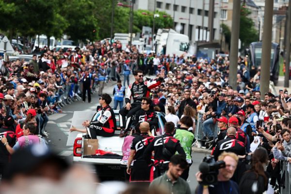 Blick auf eine Parade: Drei Rennfahrer fahren winkend auf einem Rennwagen durch ein Spalier von Fans, die rechts und links hinter Absperrungen stehen