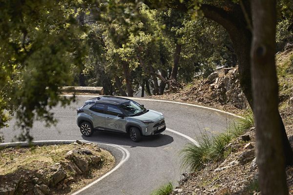 Ein hellgrauer Yaris Cross mit schwarzem Dach beim Durchfahren einer Serpentine in mediterranem Gebirge, im Vordergrund ein Baum