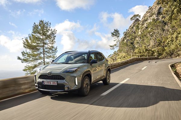 Ein hellgrauer Yaris Cross mit schwarzem Dach fährt auf einer Küstenstraße, im Hintergrund das Meer, steile Felsen und mediterrane Bäume