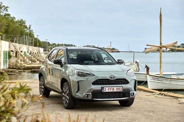 Ein hellgrauer Yaris Cross mit schwarzem Dach, geparkt an einem Anleger mit kleinen Fischerbooten, der Hafenbucht und einem Leuchtturm im Hintergrund