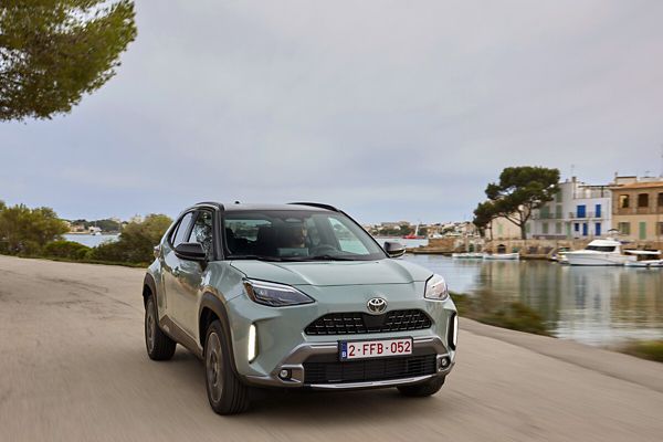 Ein hellgrauer Yaris Cross mit schwarzem Dach fährt an einem Hafenbecken entlang, im Hintergrund kleine Boote, Wohngebäude und Bäume