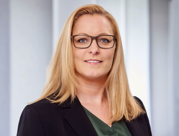 Portrait von Cathrin Koch mit langen, glatten blonden Haaren und Brille, die einen schwarzen Blazer über einem grünen Oberteil trägt