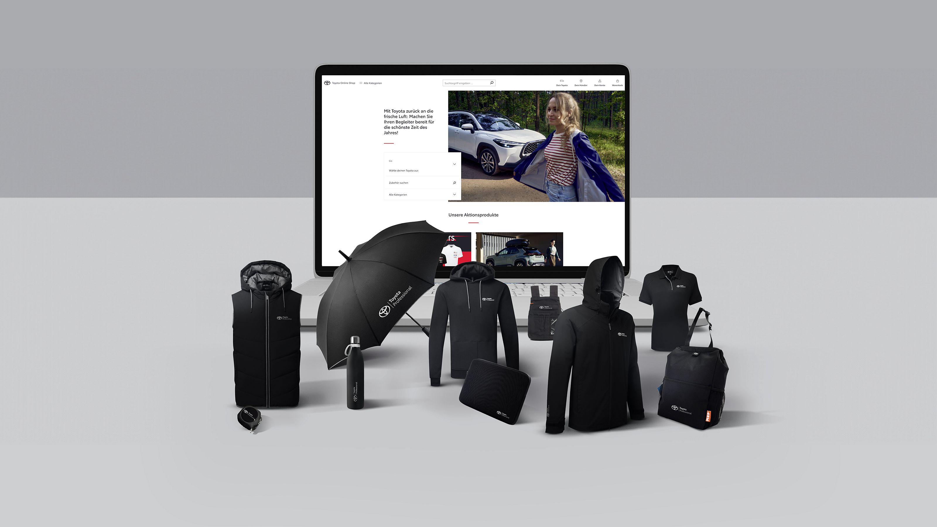 Einige schwarze Toyota-Merchandise-Artikel, darunter Schirm, Jacke und Trinkflasche sind neben einem Bildschirm angeordnet, der eine Website mit einem Toyota RAV4 im Schnee zeigt. 