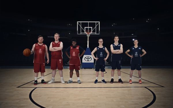 Sechs Sportler stehen in einer abgedunkelten Basketball-Halle und lächeln in die Kamera, während hinter ihnen der Basketballkorb mit erleuchtet wird.