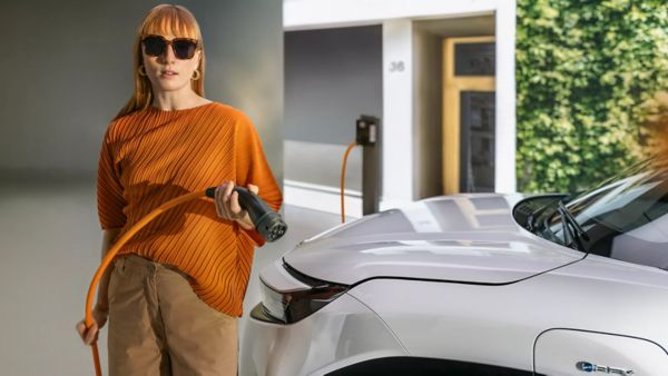 Frau mit Sonnenbrille und orangefarbenem Oberteil hält ein Ladekabel vor einem weißen Toyota Urban Cruiser – Elektromobilität trifft auf urbanen Lifestyle, Ladesituation im Alltag.