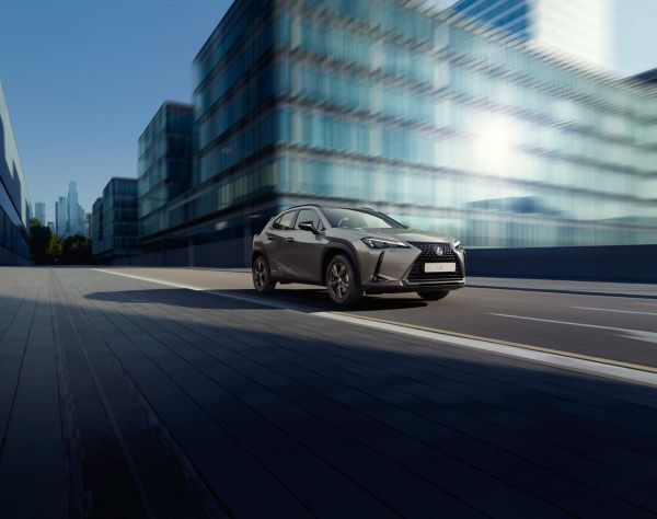 Ein silberner Lexus UX fährt auf einer modernen Stadtstraße entlang, im Hintergrund spiegeln sich Glasgebäude im Sonnenlicht.