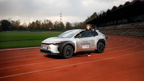 Ulrike Nasse-Meyfarth steigt in einen silbergrauen Toyota bZ4X, der auf der Laufbahn eines kleinen Stadions steht, im Hintergrund die Tribüne und Rasen