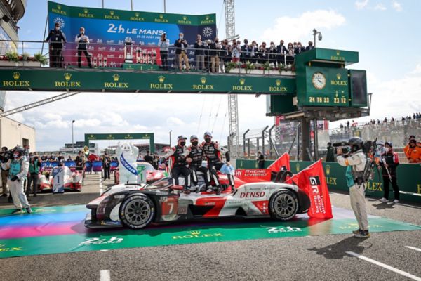 Toyotas Le-Mans-Rennwagen Nr. 7 überquert die Ziellinie, während das Team feiert. Zuschauer jubeln von den Tribünen, im Hintergrund ein Glockenturm.