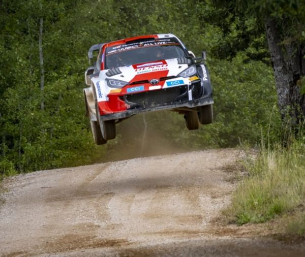 Ein Toyota Rallye-Aut, wird in der Luft eingefangen, während es eine von üppigen grünen Bäumen gesäumte Schotterstraße entlangfährt. 