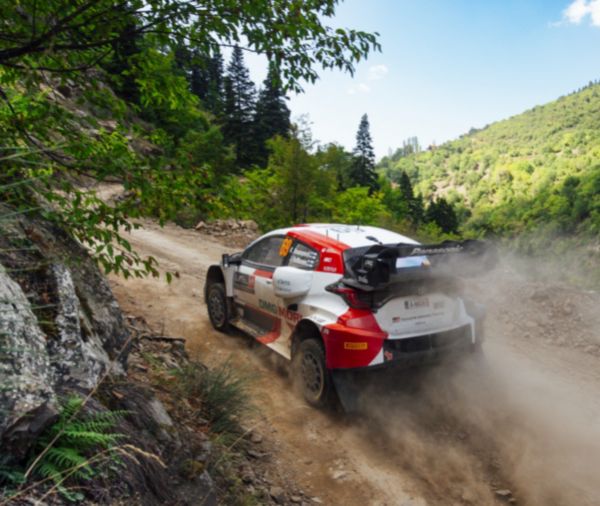 Ein Toyota Rallye-Auto navigiert eine staubige Schotterstraße, umgeben von üppigen grünen Hügeln und Bäumen. 