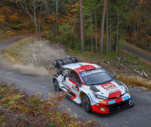 Ein Rallye-Auto in rot-weißem Design navigiert eine kurvenreiche Waldstraße entlang und wirbelt Staub auf, während es durch buntes Herbstlaub rast.