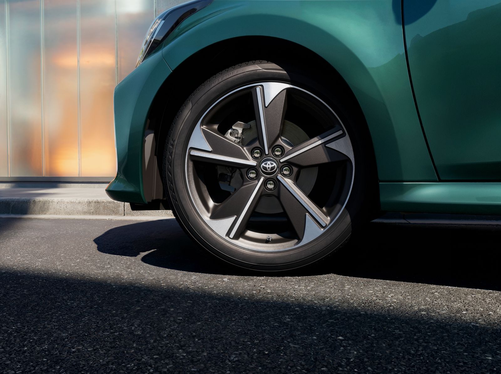 Eine junge Frau lehnt am hinteren Kotflügel eines dunklen Yaris GR Sport, der vor einem von der Sonne beschienenen Laubbaum steht
