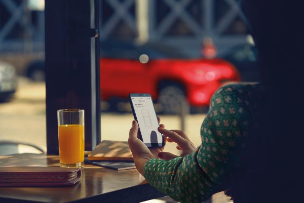 Žena sedí v kavárně. Zatímco používá aplikaci MyToyota na telefonu, na pozadí stojí zaparkována její Toyota Yaris Cross.