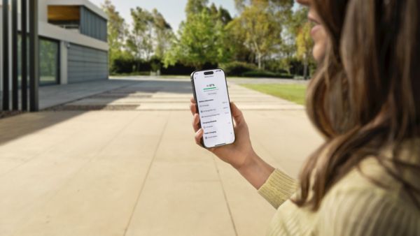 Nő áll egy modern ház előtt, okostelefont tart a kezében, amelyen egy elektromos autó töltését felügyelő alkalmazás látható.