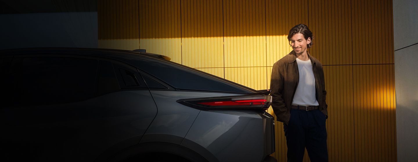 Ein Mann steht lächelnd neben dem Heck eines modernen Autos vor einer gelb beleuchteten Wand; links im Bild sind teilweise Werbetexte und ein Button sichtbar.