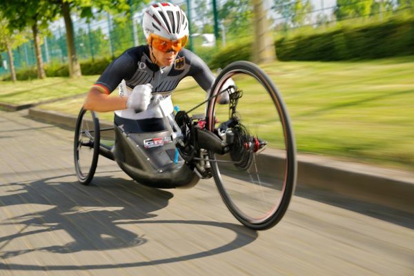 Paralympics-Sportlerin während der Fahrt in ihrem Handbike schräg von vorne dynamisch fotografiert, im Hintergrund Bäume und ein Sportgelände