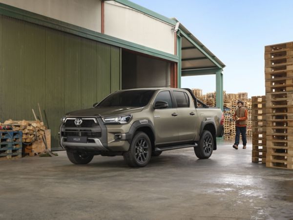 Ein Mann in Arbeitskleidung nähert sich einem vor einer Lagerhalle stehenden grünen Hilux, im Hintergrund hohe Stapel mit Holzpaletten