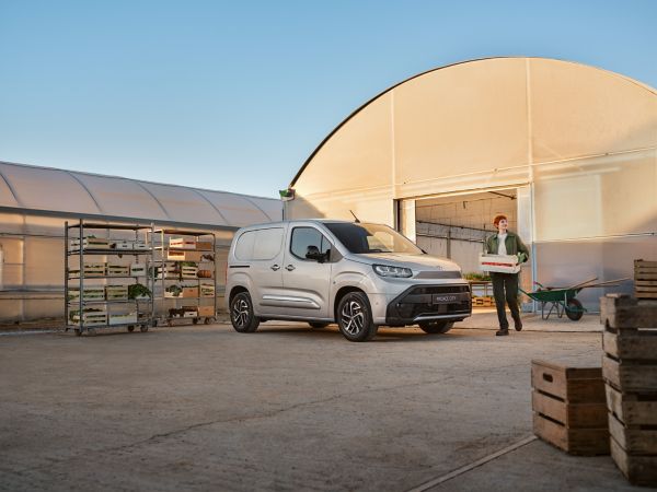 Eine junge Frau in Arbeitskleidung trägt eine Gemüsekiste aus einem Gewächshaus zu einem davor geparkten grauen Proace City.