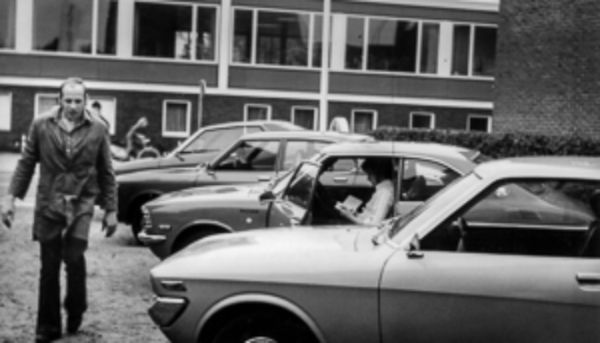 Ein Schwarzweiß-Bild von einem Mann, der an geparkten Fahrzeugen vorbeiläuft 