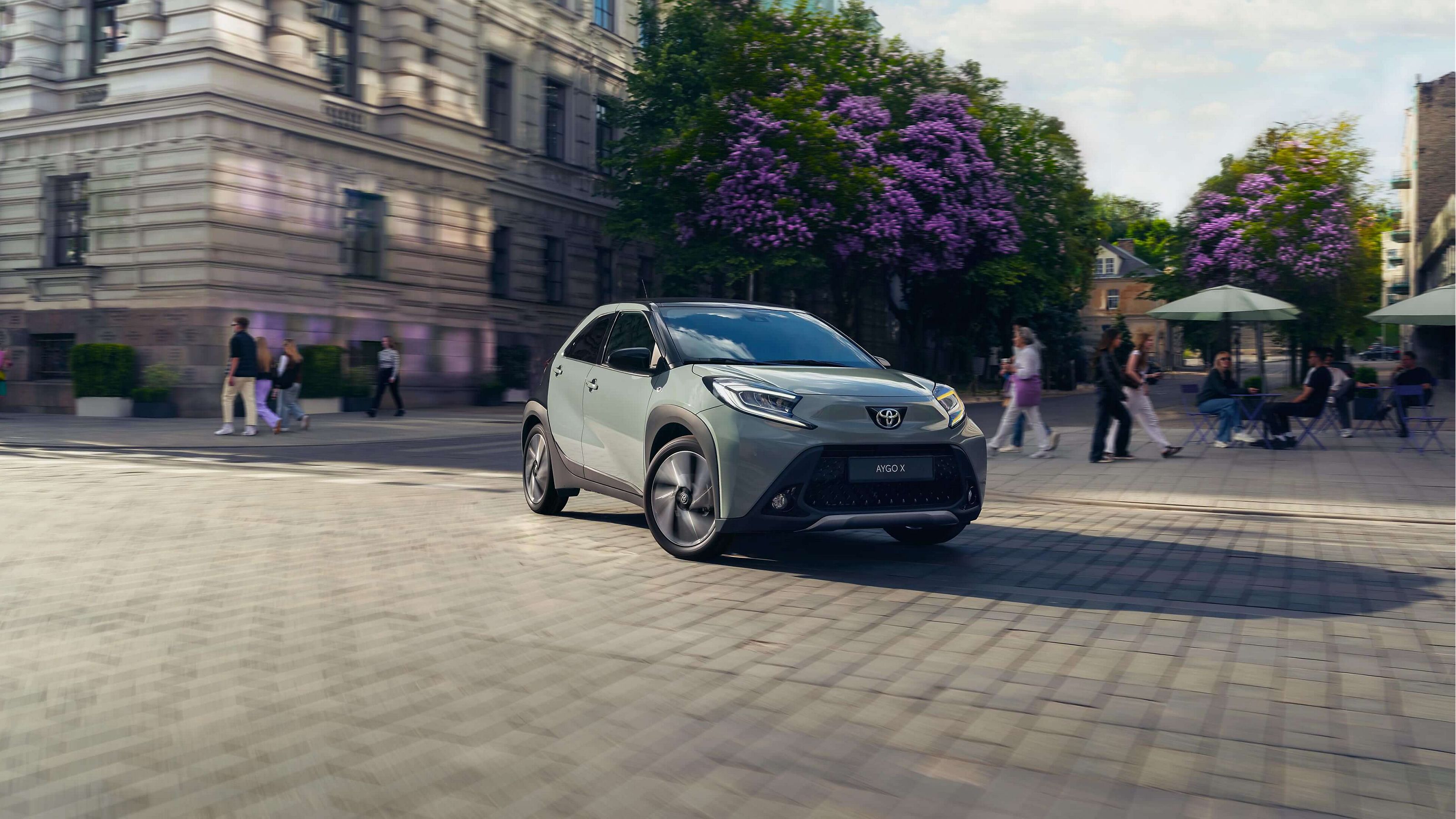 Ein Toyota Aygo X in einer städtischen Umgebung, im Hintergrund sind Bäume und Menschen zu sehen
