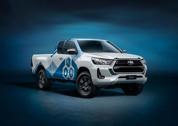 Ein weißer Toyota Hilux Pick-up mit blauer Bedruckung und drei Symbolen für Wasserstoff, Wasser und Brennstoffzelle auf der Tür, blaue Studio-Umgebung