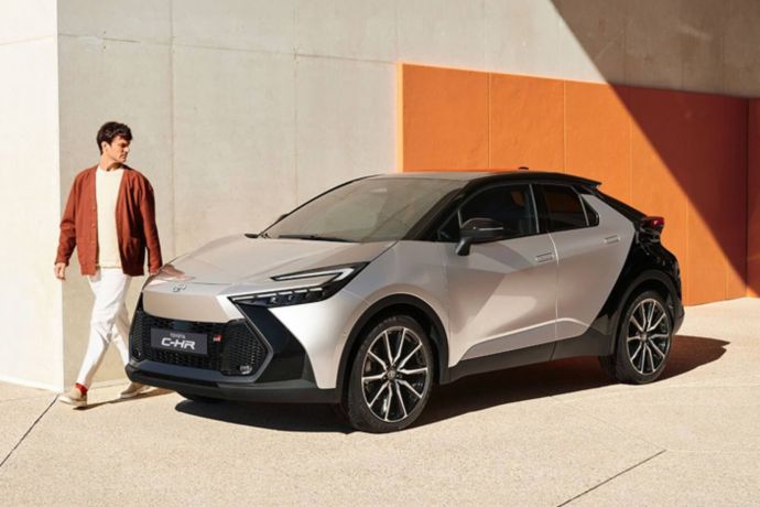 Person läuft an der Seite eines silbernen Toyota Kompakt-SUVs entlang, das vor einer Orange-beigen Wand geparkt ist.