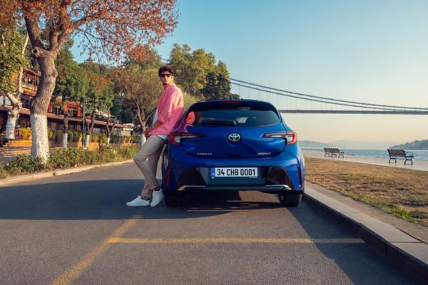 yolda duran mavi Corolla Hatchback Hybrid yanında araca yaslanan bir adam