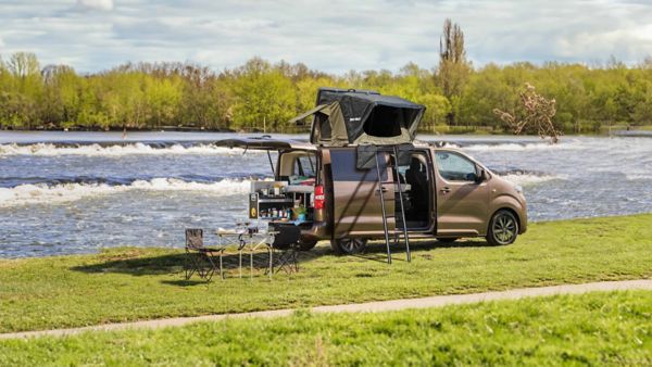 Ein am Fluss geparktes Wohnmobil mit geöffneter Seitentür, die den Blick auf Stauraum und Gartenmöbel freigibt, die auf dem Rasen aufgestellt sind.