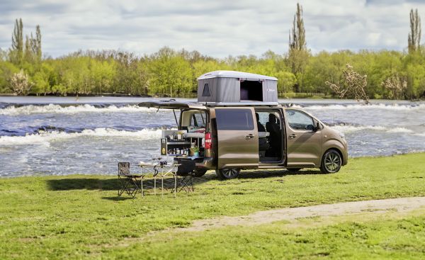 Ein am Fluss geparktes Wohnmobil mit geöffneter Seitentür, die den Blick auf Stauraum und Gartenmöbel freigibt, die auf dem Rasen aufgestellt sind.