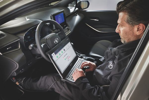 Ein Techniker sitzt in einem Auto und verwendet einen Laptop, während im Hintergrund ein Armaturenbrett zu sehen ist.