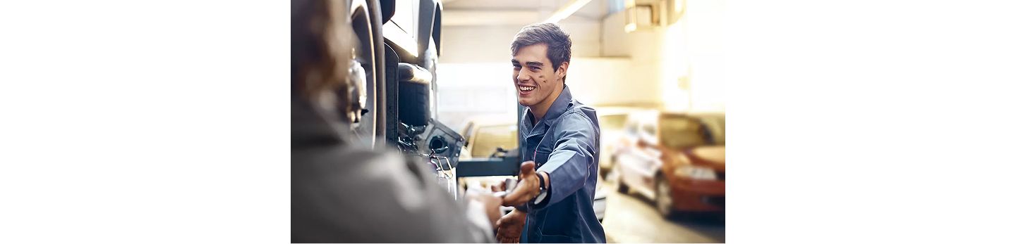 Eine Person in Arbeitskleidung reicht einem anderen Menschen ein Werkzeug in einer Werkstatt, im Hintergrund sind Autos und Werkstattausrüstung sichtbar.