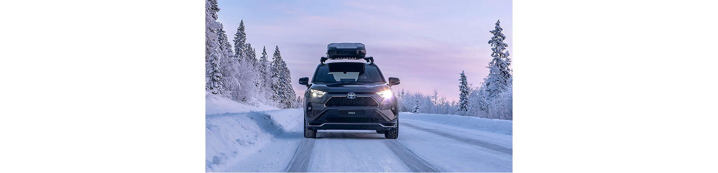 Ein schwarzer SUV mit Dachbox fährt auf einer verschneiten Straße, umgeben von schneebedeckten Bäumen unter einem rosa-blauen Winterhimmel.