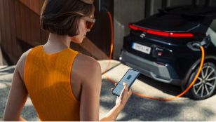 Ein schwarzer Toyota C-HR wird per Wallbox geladen, während eine Frau in der App den Batteriestand checkt