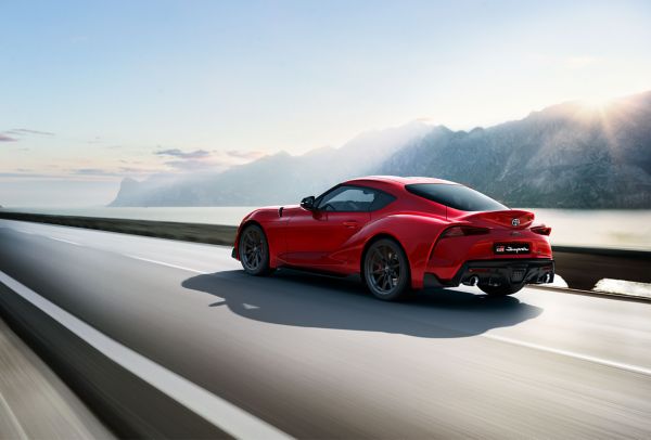 Ein roter Sportwagen, der Toyota Supra, fährt über eine schnell vorbeiziehende Straße, während im Hintergrund ein Bergpanorama von der Sonne angestrahlt wird.