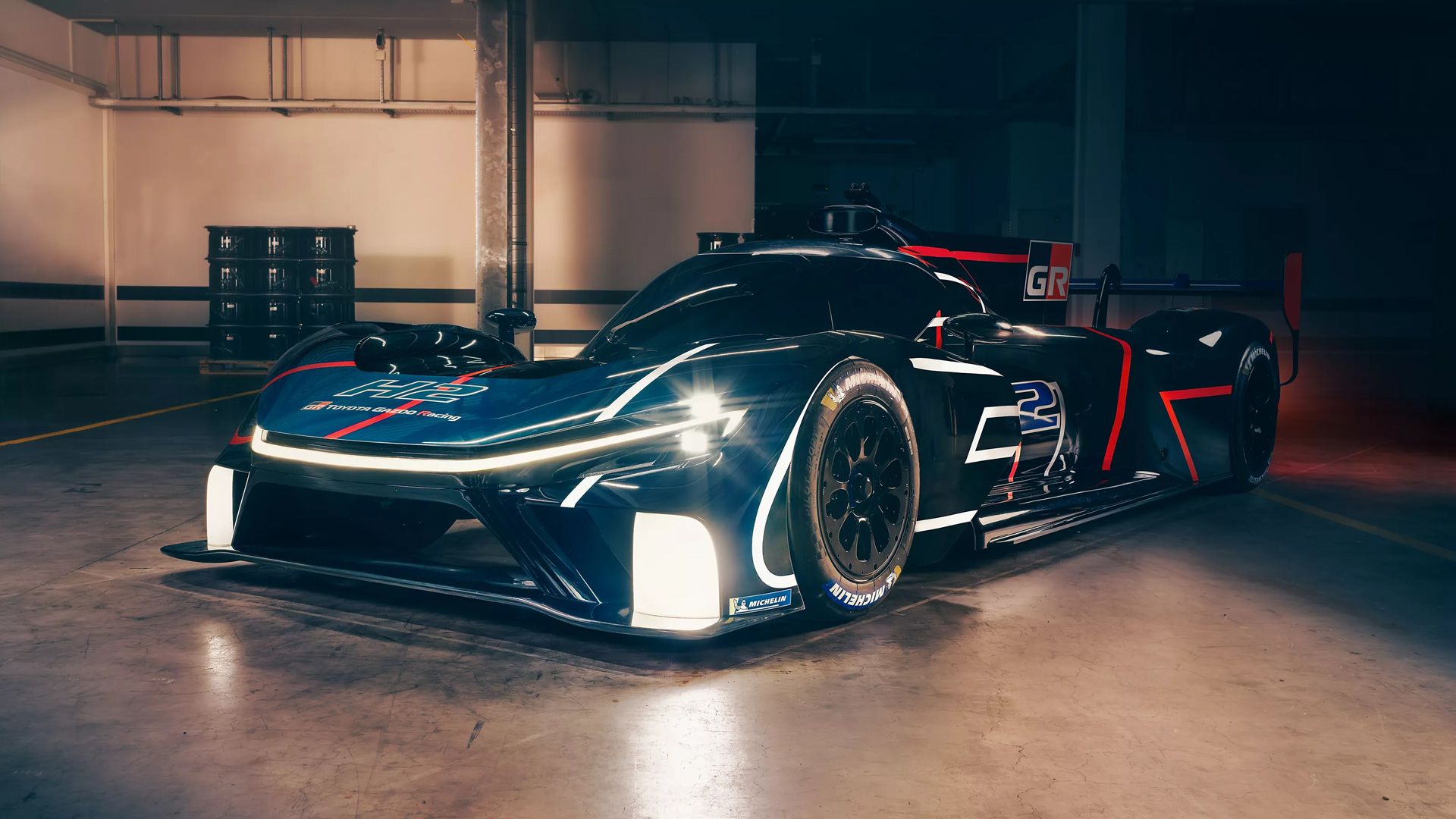 Egy sötétkék Toyota Gazoo Racing versenyautó, fehér és piros mintákkal, kivilágított fényszórókkal, egy garázsban állva.