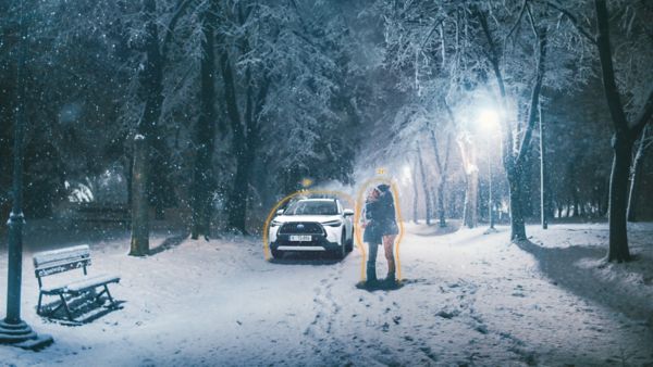 Ein Paar steht nachts in einem verschneiten Park dicht beieinander, beleuchtet von Straßenlaternen, während im Hintergrund ein geparktes Auto zu sehen ist.