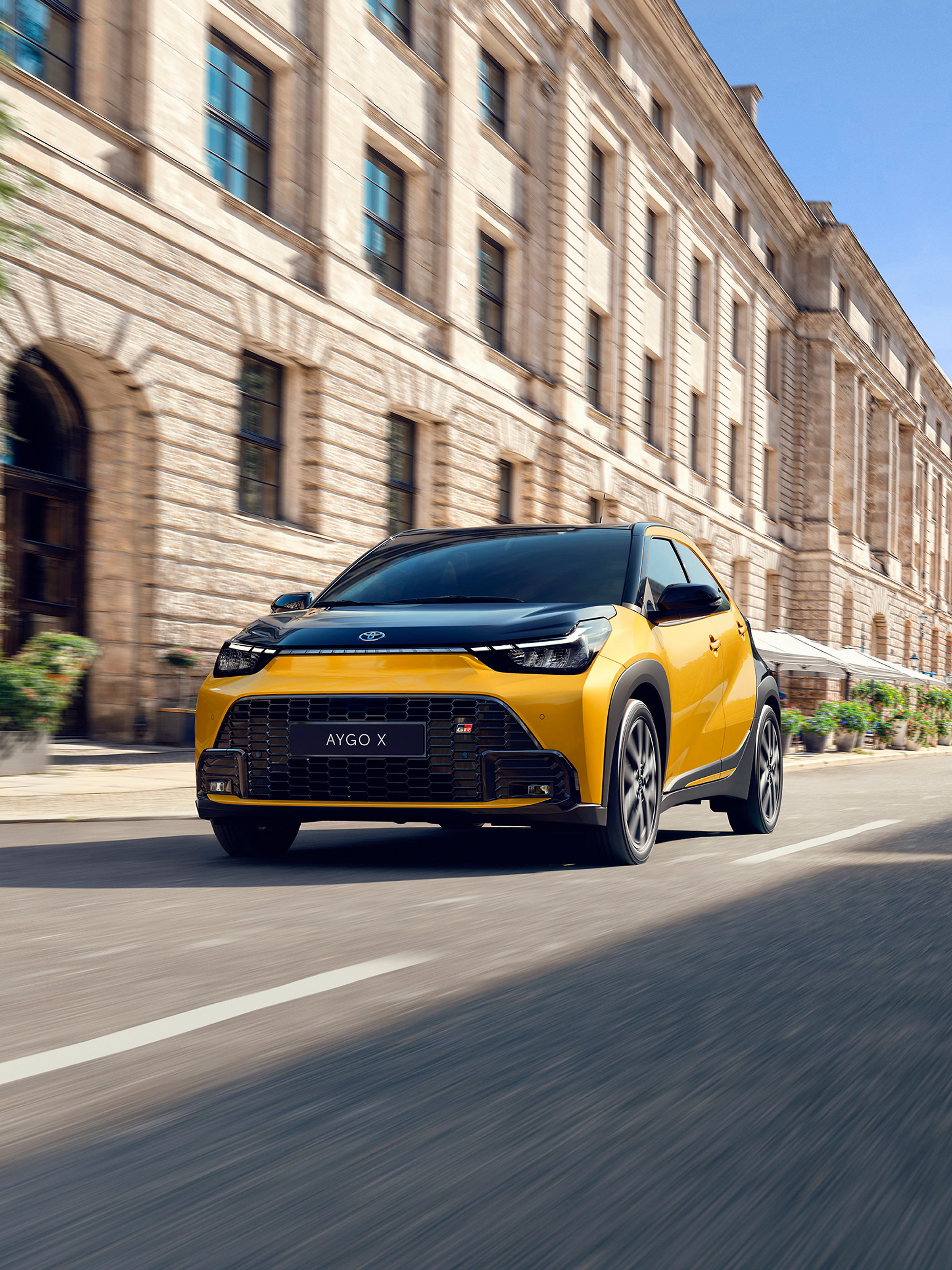 Gelber Toyota Aygo X GR Sport fährt auf einer Stadtstraße entlang historischer Gebäude; Aufnahme aus niedriger Perspektive mit Bewegungsunschärfe im Hintergrund.