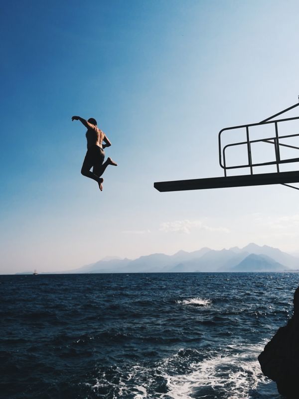 Eine Person in der Luft, die von einem Sprungturm in ein strahlend blaues Meer springt, mit Bergen im Hintergrund unter einem klaren Himmel