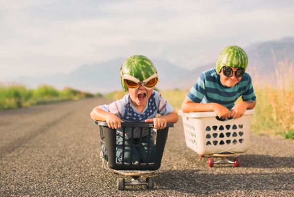 Zwei Kinder rasen in verspielten Go-Karts, die aus einem Einkaufskorb und einem Wäschekorb gebaut wurden, eine Schotterstraße hinunter. Sie tragen Helme aus Wassermelonenschalen und übergroße Sonnenbrillen.