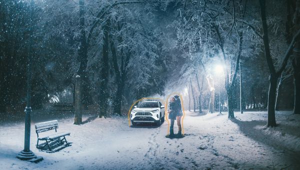 Zwei Menschen im Schnee unter Bäumen sich umarmend. Im Hintergrund ein Toyota. Es ist dunkel, Laternen leuchten.