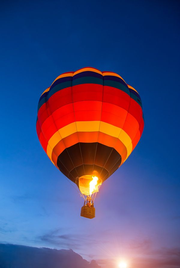 Ein farbenfroher Heißluftballon mit roten, orangefarbenen und gelben Streifen wird beim Aufsteigen gegen den Dämmerungshimmel beleuchtet.