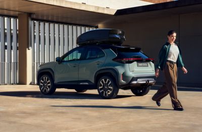 Hellgrüner Toyota Yaris Cross mit schwarzer Dachbox steht in moderner Architekturkulisse, während eine Frau in eleganter Freizeitkleidung daran vorbeigeht.