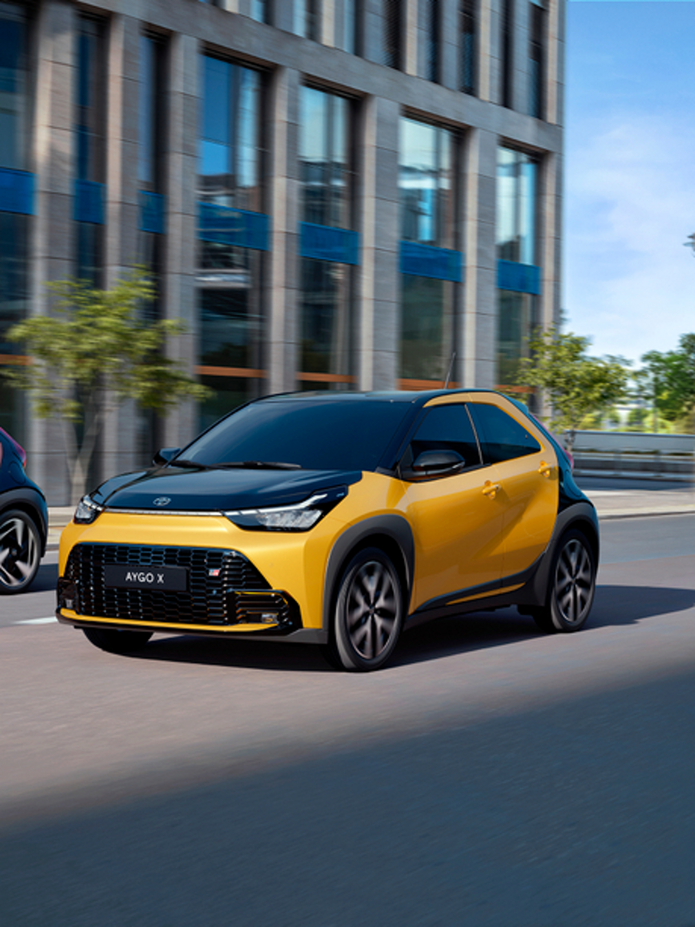 Zwei Toyota Aygo X fahren durch eine Stadtstraße vor moderner Büroarchitektur in urbaner Umgebung.