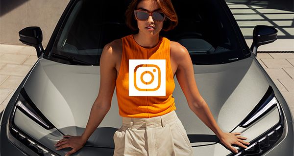 Nő narancssárga felsőben és napszemüvegben, egy világos színű Toyota motorháztetőjére támaszkodva.