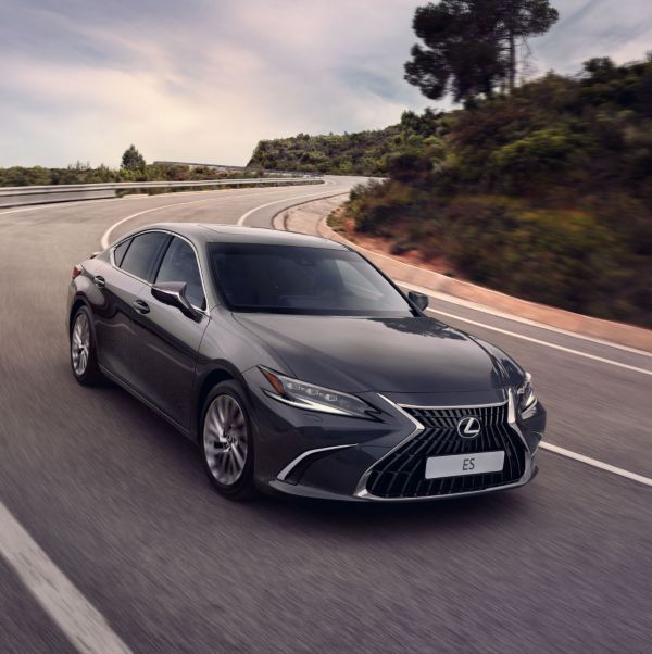 Ein eleganter grauer Lexus ES 300h fährt auf einer kurvigen Straße, umgeben von Grün unter einem teilweise bewölkten Himmel.