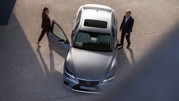 Zwei Personen beschäftigen sich mit einem silbernen Lexus LS, der auf einer gepflasterten Fläche geparkt ist. Eine Person steht an der offenen Fahrertür, die andere nähert sich von vorne.